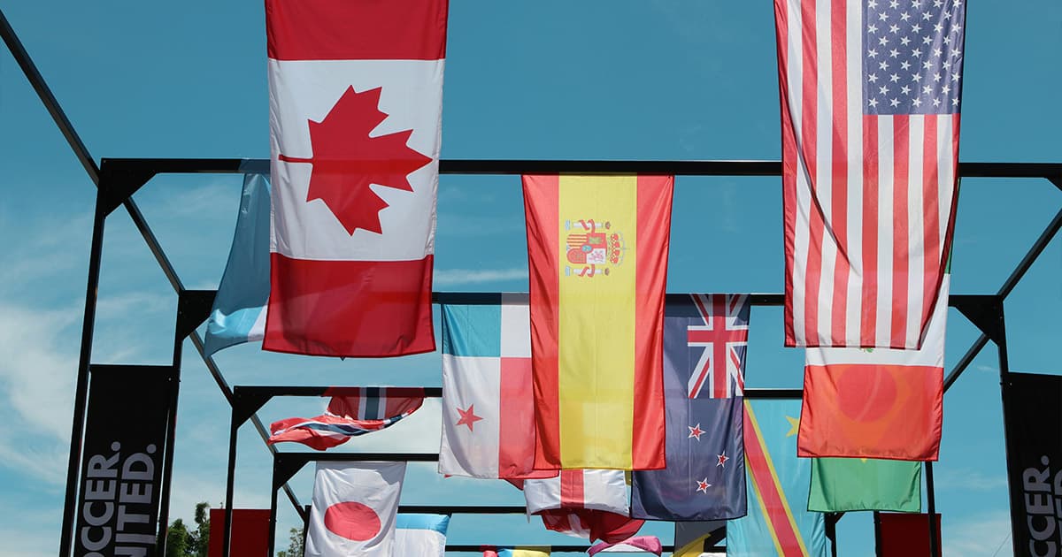 Several national flags, including those of Canada, the United States, Spain, and the United Kingdom, hang together outdoors against a clear blue sky.