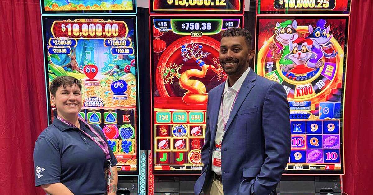 Two people stand smiling in front of colorful slot machines at a casino or gaming event, with bright screens showing jackpots and cartoon graphics behind them.
