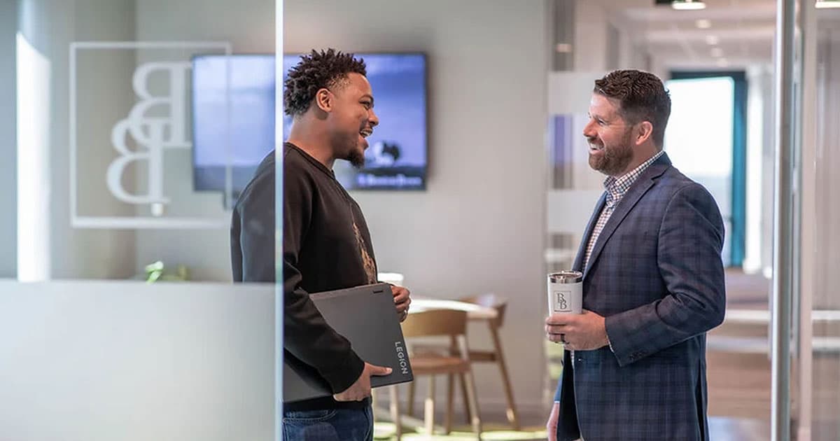 Two men stand in an office hallway, smiling and talking. One holds a laptop, the other holds a travel mug. Glass walls and office furniture are visible in the background.