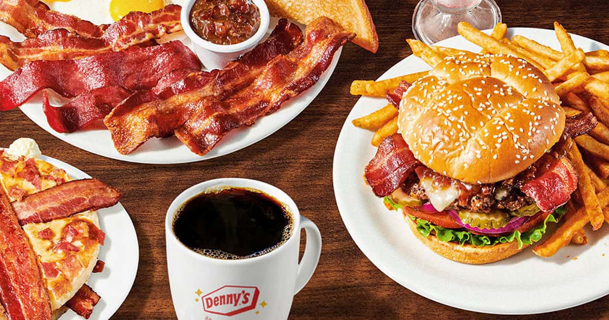 Plates with bacon, eggs, toast, fries, a bacon cheeseburger, pizza slices with bacon, a cup of coffee, and a small bowl of salsa on a wooden table.