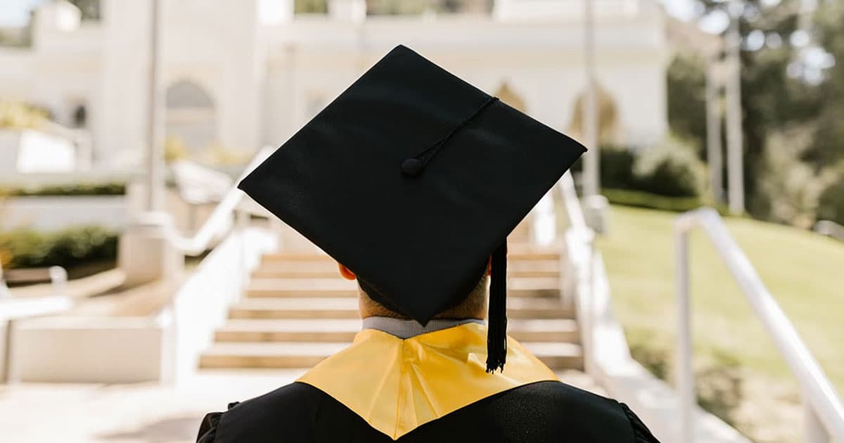 A person wearing a graduation cap and gown stands outdoors, facing a staircase and a blurred building in the background.