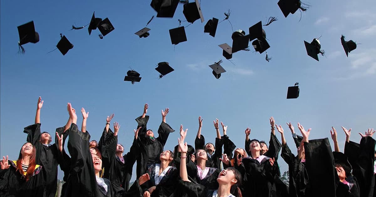 A group of graduates in caps and gowns cheer and throw their caps into the air under a clear blue sky, celebrating their graduation.