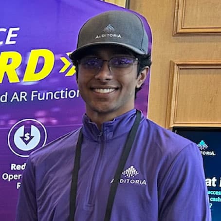 Smiling person in glasses and a gray hat, wearing a purple jacket with the Auditoria logo, stands in front of a purple and yellow display booth.