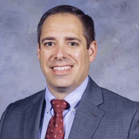 A man in a gray suit, light blue shirt, and red patterned tie smiles in front of a gray mottled background.