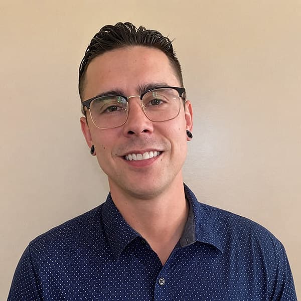 Smiling man with glasses, short dark hair, and black earrings, wearing a dark blue collared shirt with a subtle dotted pattern, standing in front of a beige wall.
