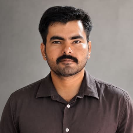 A man with short black hair, a mustache, and beard, wearing a dark brown collared shirt, stands against a plain gray background, looking directly at the camera with a neutral expression.