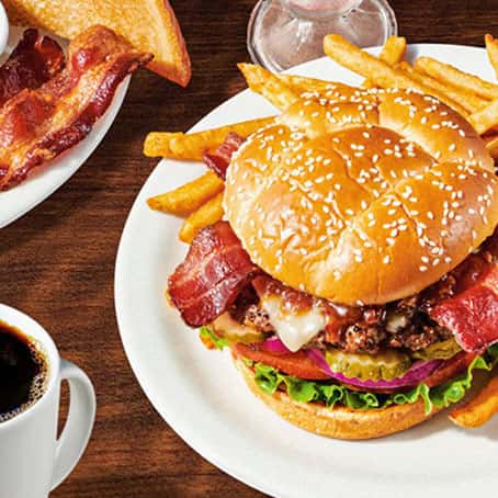 A plate with a bacon cheeseburger, fries, and a cup of black coffee on a wooden table. The burger has lettuce, tomato, pickles, onions, cheese, and bacon on a sesame seed bun. Bacon and toast are on a separate plate.
