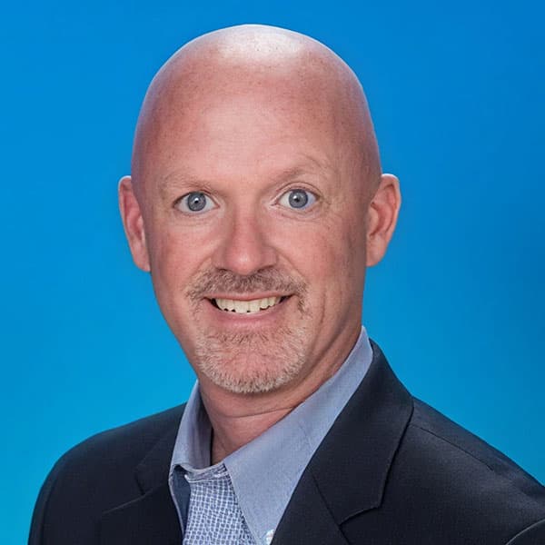 A smiling bald man with blue eyes, a goatee, and light skin wearing a black blazer and light blue collared shirt, posed against a solid blue background.