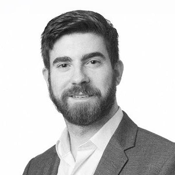 A man with a beard and short hair, wearing a suit jacket and white shirt, poses for a professional black-and-white headshot against a plain background.