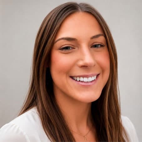 A woman with long brown hair and a white top smiles in front of a plain light background.