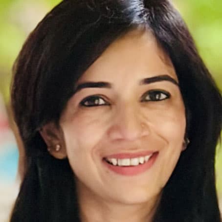 A woman with dark hair and brown eyes smiles at the camera. She is wearing small earrings and has a blurred green background behind her.