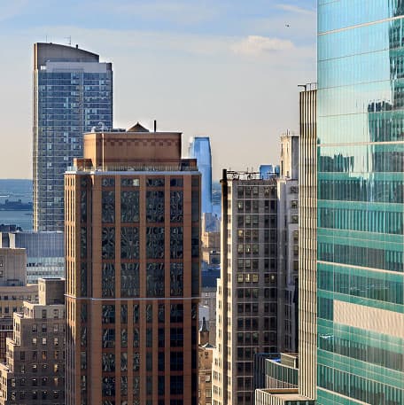 Sunlight reflects off the glass and concrete facades of several tall, modern office buildings in a city skyline under a clear blue sky.