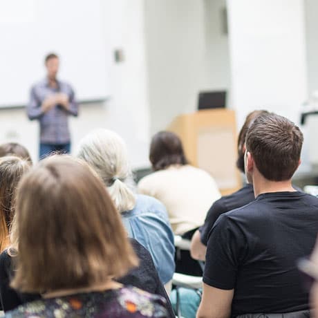 A group of people sit facing a speaker at the front of a classroom or seminar room. The letters FPO in large pink text are overlaid on the image.