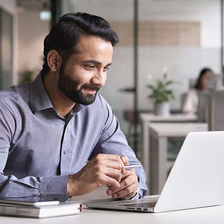 A man wearing glasses and a sweater types on a laptop in a modern office setting with glass walls and blurred colleagues in the background.