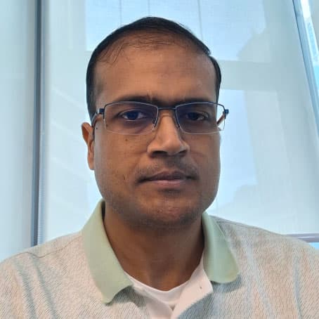 A man with short hair and glasses wearing a light-colored collared shirt sits indoors in front of a window with natural light.