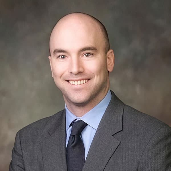 A smiling bald man in a gray suit, blue shirt, and dark tie poses in front of a blurred brown and gray background.