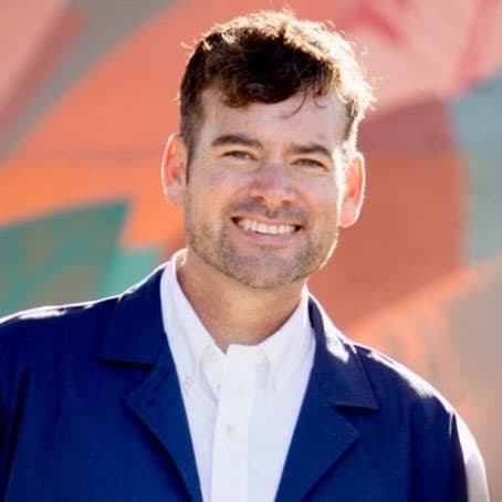 A man with short brown hair and light stubble smiles at the camera, wearing a white shirt and a blue jacket. The background is blurred with colorful, abstract shapes.