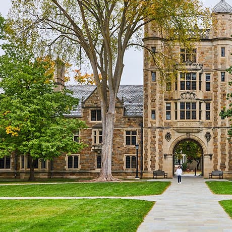 A person walks on a path toward a historic stone university building with an arched entrance, tall windows, and trees with green and yellow leaves in the foreground.
