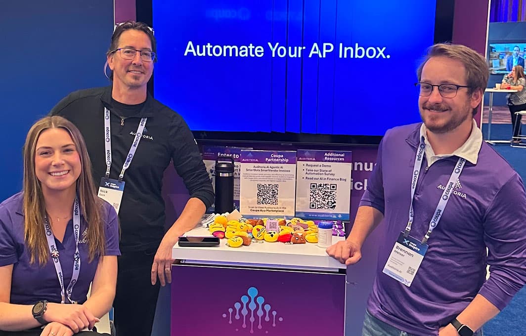 Three smiling people at a booth with a display reading Automate Your AP Inbox. They wear name badges and purple shirts. The table has rubber ducks, QR codes, and promotional items.