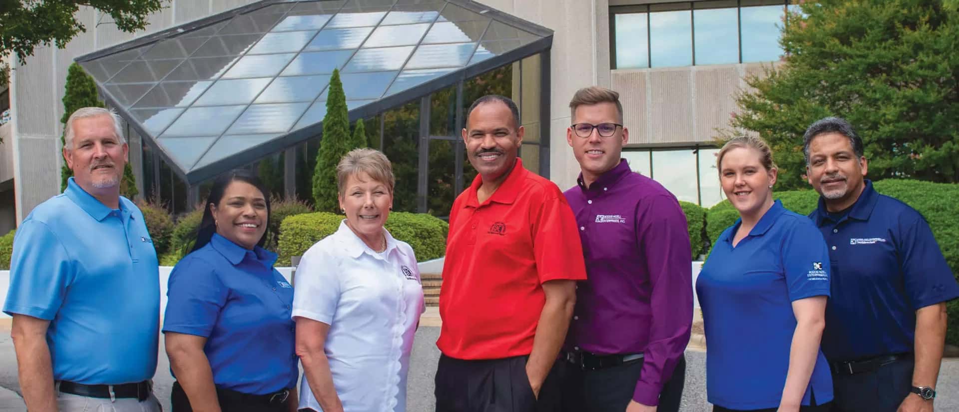Seven people stand outdoors in front of a modern building, smiling at the camera. They wear collared shirts in different colors, possibly representing a team or organization. Greenery surrounds the building entrance behind them.