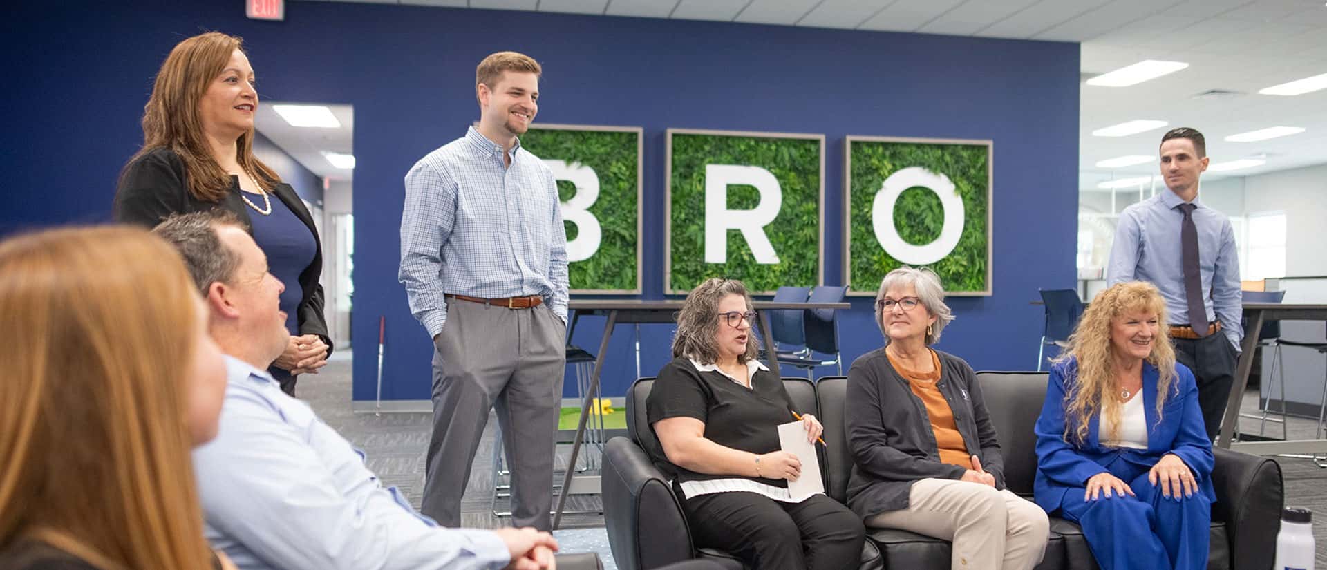 A group of professionally dressed people sit and stand in an office lounge area, smiling and talking. Behind them, three wall panels with the letters B, R, and O are visible.