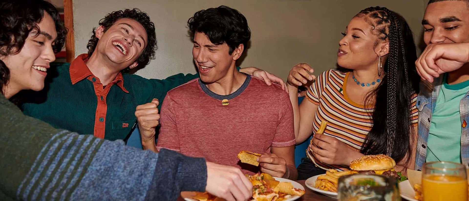 Five friends sit around a table laughing and eating together, enjoying burgers and fries in a casual, cheerful setting.