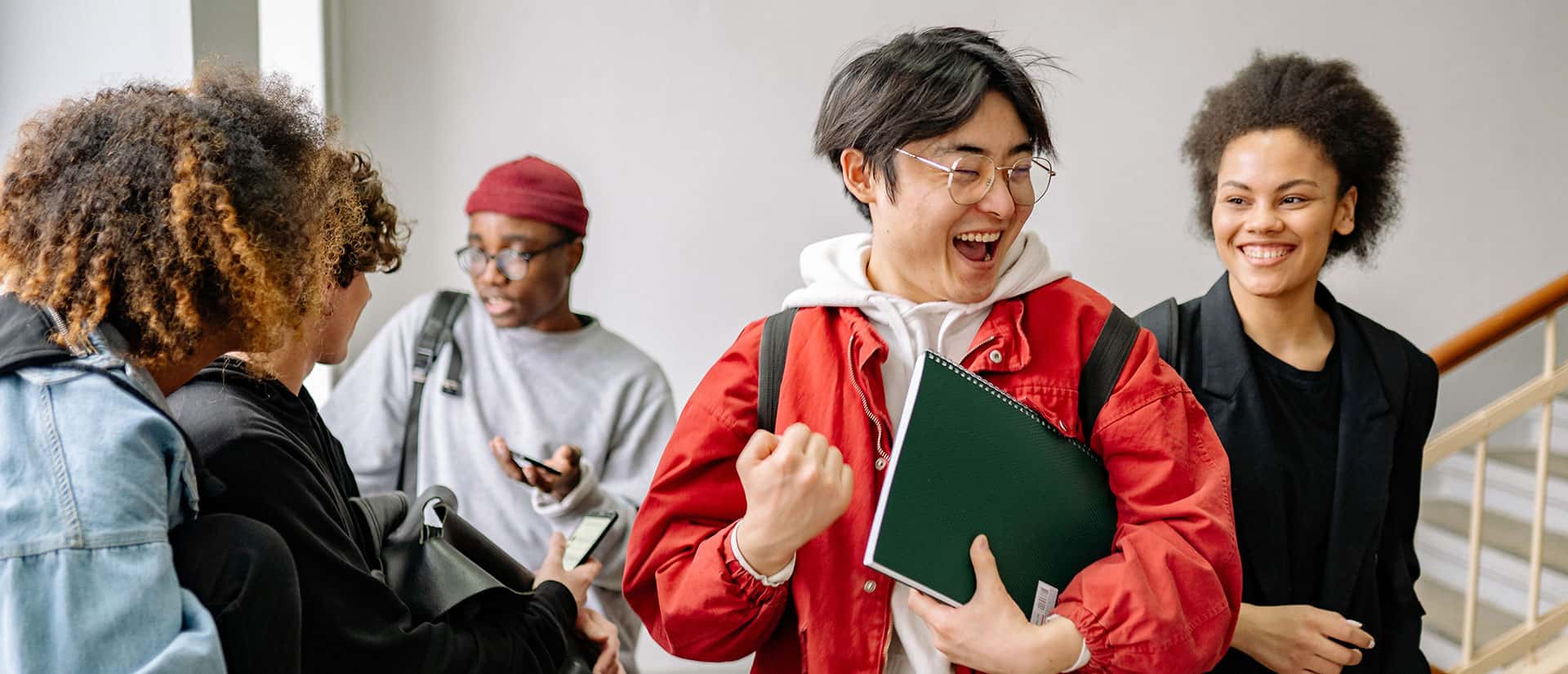 Four students are talking and laughing together indoors. One person in a red jacket is smiling and holding a notebook, while others stand nearby with backpacks, looking happy and engaged.