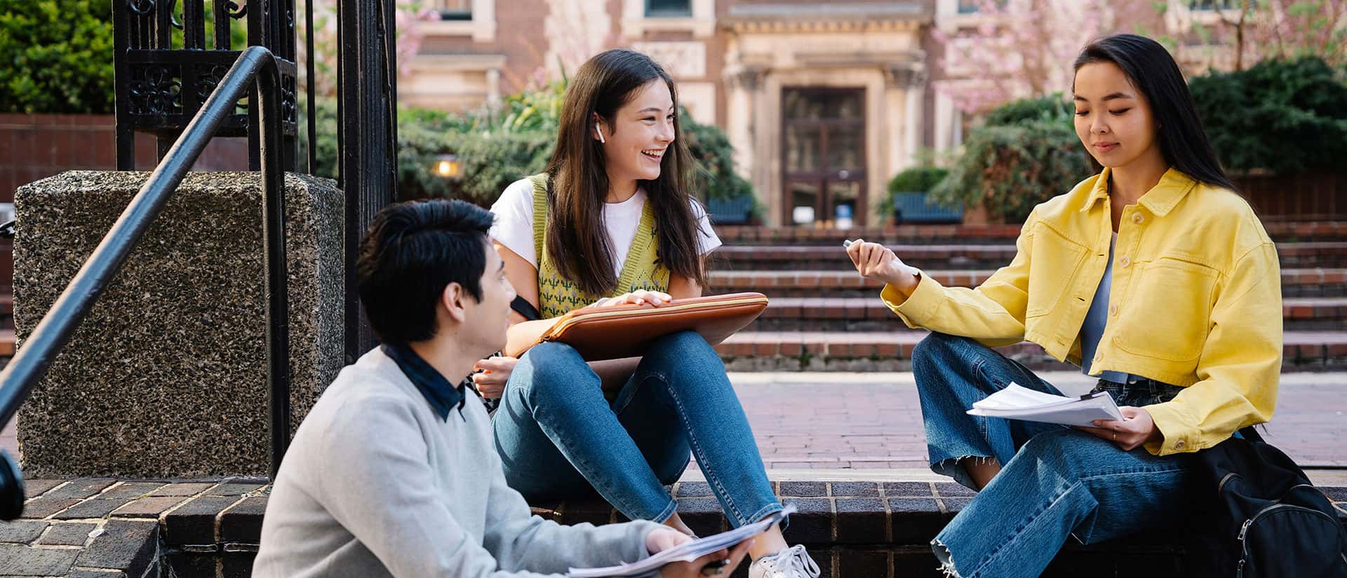 Three students sit on outdoor steps, talking and studying together with notebooks in hand. Two are sitting on a step, and one is sitting lower down, engaged in the conversation. Trees and a building are in the background.