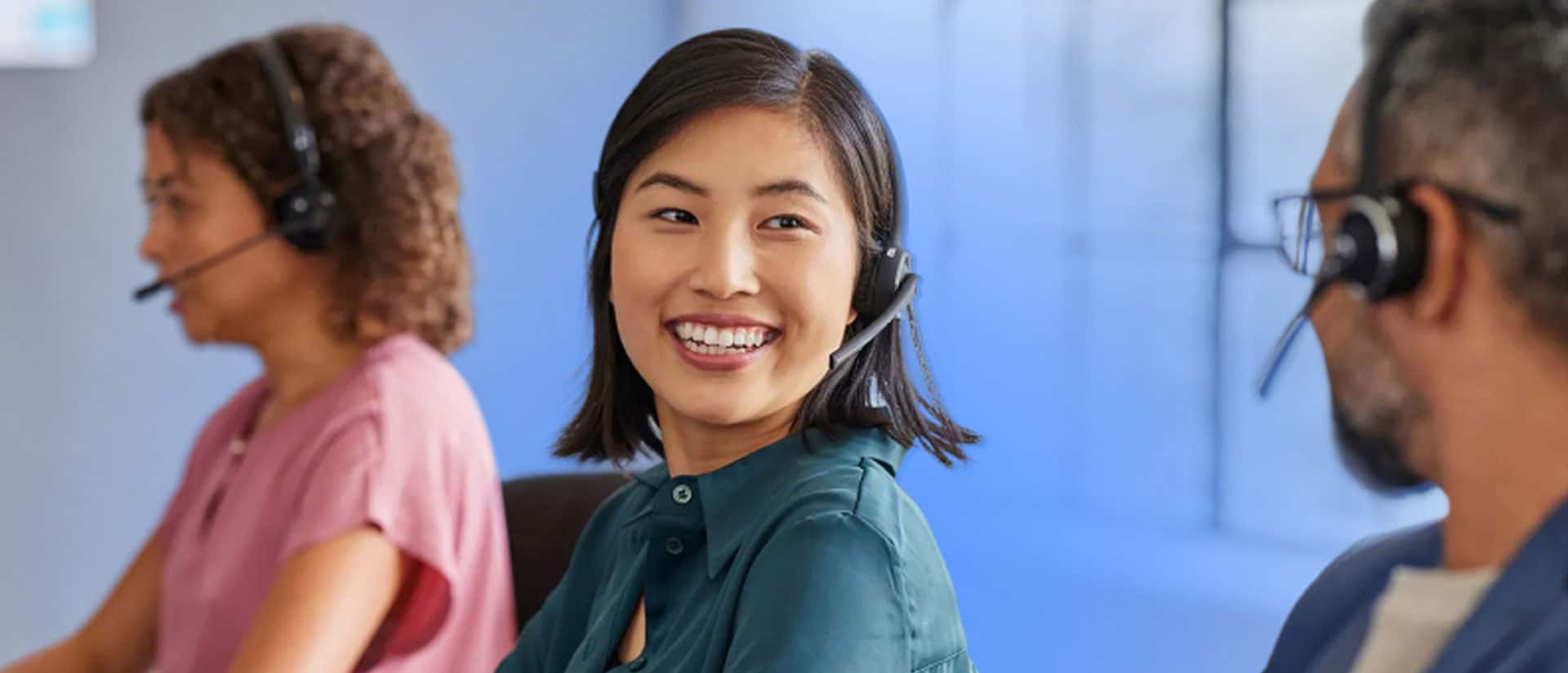 Three people wearing headsets sit at desks in a modern office. The woman in the center, smiling, turns to speak with the man beside her. The overall atmosphere is friendly and professional.