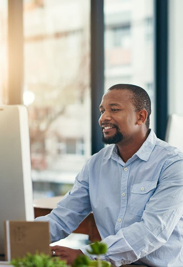 A man in a light blue shirt sits at a desk, smiling while working on a computer in a bright office. FPO is written in pink over the image.