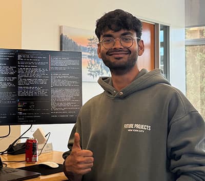 A person wearing glasses and a Future Projects New York City hoodie smiles and gives a thumbs up while standing next to a monitor displaying code. A can of soda is on the desk.