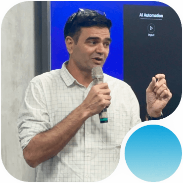 A man in a light plaid shirt speaks into a microphone and gestures with his hand in front of a screen displaying AI Automation text.