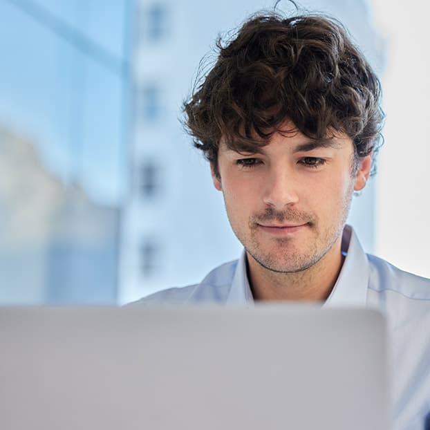 A young man with curly dark hair focuses on a laptop in a bright, modern office, wearing a light blue shirt; his expression is calm and attentive.
