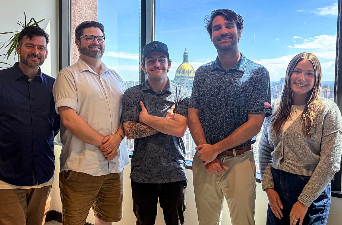 Five people stand smiling in an office with large windows, showing a city skyline and a gold-domed building under a blue sky in the background.