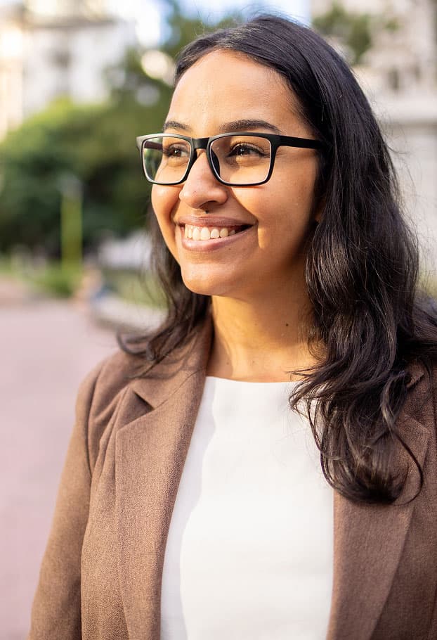 A woman wearing glasses and a brown blazer smiles outdoors, holding a tablet. Green trees and blurred buildings are in the background.